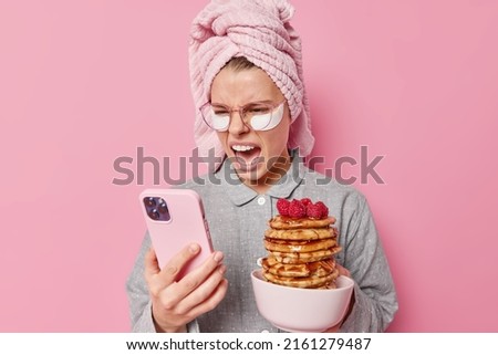 Irritated young woman yells angrily focused at smartphone scrolls newsfeed wears towel on head and slumber suit prepared appetizing pancakes with rasberry for family breakfast feels outraged Royalty-Free Stock Photo #2161279487