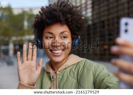 Cheerful curly woman takes self portrait via smartphone waves palm says hello listens music via headphones smiles gladfully poses outdoors against blurred background has recreation using technology
