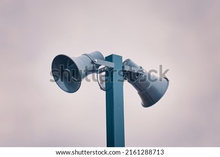 Hazard warning system. Tall metal column with two gray loudspeakers against cloudy sky. Providing security in the city, notification of emergencies. Copy space for text. Royalty-Free Stock Photo #2161288713