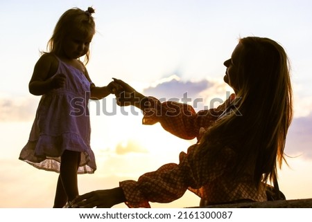Silhouettes of young happy mother with little girl, cute toddler daughter in lace dress against a sky background. Mom supporting child, toddler. Kids protection. Children's Day June 1. Family welfare.