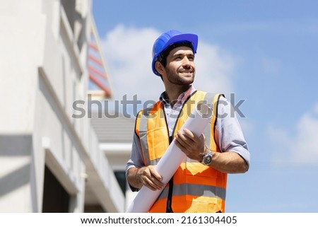 A smiling caucasian builder in a white hard hat and yellow fluorescent jacket holds clipboard, stands on the roof, inspects construction site and makes notes. Royalty-Free Stock Photo #2161304405