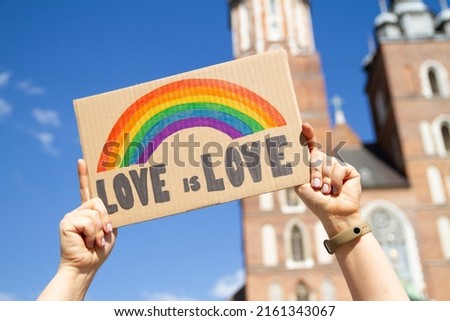 Love is Love rainbow flag placard sign, symbol of LGBT love. Pride Parade equality march in Krakow, Poland to support and celebrate LGBT+, LGBTQ. Saint Mary's Basilica (Mariacki Church Kraków) in bg.