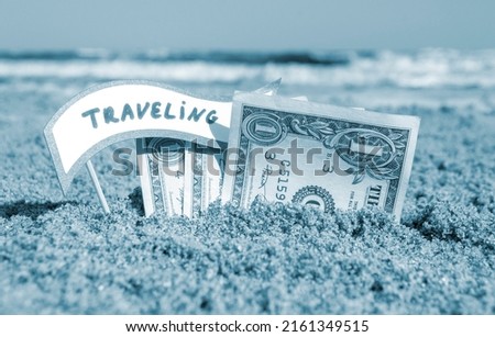 Paper bills one dollar and small stick with paper speech bubble with word Traveling buried in sand beach background sea sunny summer day. Concept Money travel tourism vacation holiday relax Blue color