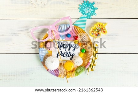bright multicolored party accessories, sweets, marshmallows and paper dish with birthday invitation on light wooden pastel background