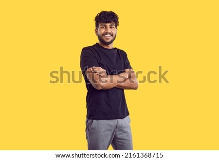 Studio shot of happy young man in casual wear. Handsome Indian guy with modern haircut in black Tshirt standing with his arms folded isolated on yellow background, looking at camera and smiling