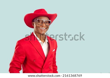 Portrait of happy extravagant dark skinned showman on pastel light blue background. African American man in red jacket, cowboy hat and shiny glasses smiles with snow-white smile near copy space.