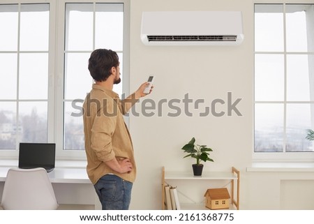 Man with remote control from air conditioner creates comfortable temperature for himself. Young man turns on air conditioner or adjusts mode for air conditioning house. Climate system concept. Royalty-Free Stock Photo #2161368775