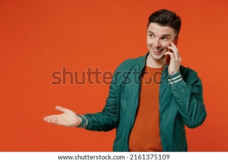 Smiling happy cheery young brunet man 20s wears red t-shirt green jacket talk speak on mobile cell phone conducting pleasant conversation look aside isolated on plain orange background studio portrait