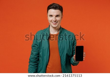Smiling charismatic happy young brunet man 20s he wears red t-shirt green jacket hold in hand use mobile cell phone with blank screen workspace area isolated on plain orange background studio portrait