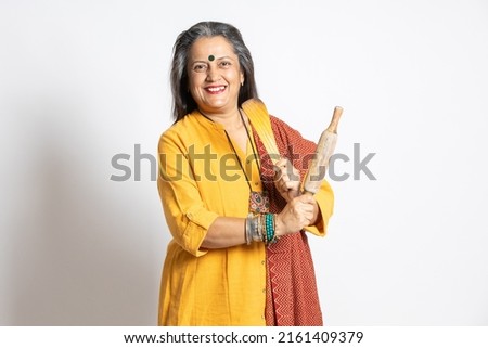Portrait of happy mature senior indian woman holding rolling pin and wooden spoon isolated on white background. home maker, kitchen concept. Royalty-Free Stock Photo #2161409379