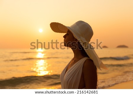 Young beautiful girl in a white dress and straw hat on a tropical beach at sunset. Summer vacation concept.