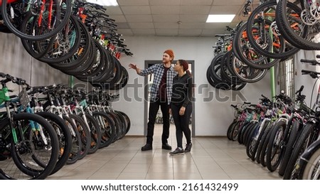 Caucasian male seller showing bicycles for focused african american female customer in modern bike shop. Set of variety bikes for extreme sport, urban lifestyle and children. Shopping concept