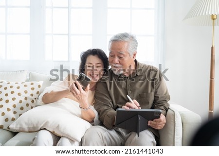 Asian senior couple in living room at home.Wife browsing online on smartphone showing something to her husband while husband is also using a tablet. Royalty-Free Stock Photo #2161441043