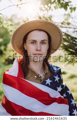 Smiling young caucasian woman in beige hat wrapped in big usa United States flag standing on background mountains at sunny day. Hipster girl feeling freedom, independence concept. July 4th concept