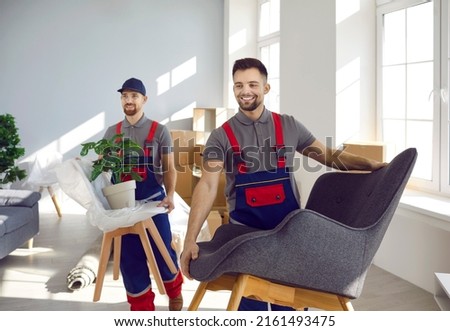 Team of happy workers from truck delivery and moving service company together removing furniture and other stuff from apartment. Two strong men in uniforms carrying chairs, armchairs and house plants Royalty-Free Stock Photo #2161493475