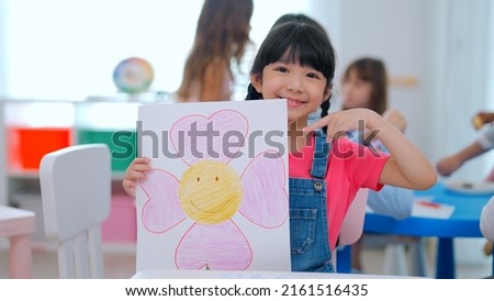 Portrait of adorable girl showing piece of work to camera drawing and painting picture on paper to teacher taking video camera with beautiful smile enjoying in the classroom, Learning with fun concept