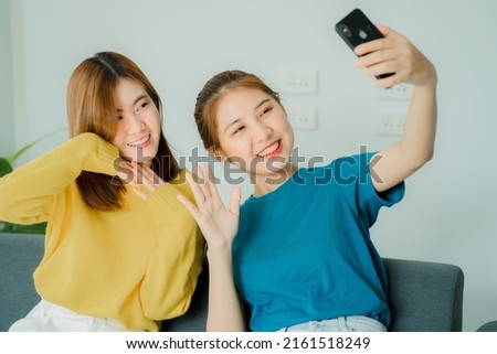 Two Asian women sit on gray sofas with smartphones taking pictures on social media in their living room at home.