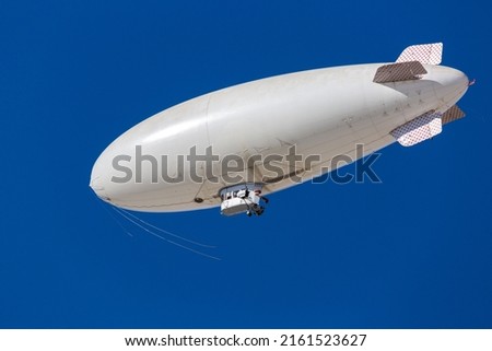 A white blimp, airship, or dirigible flying in blue sky. Close up detail of an unmarked zeppelin like flying vehicle. Floating high above in clear skies. Room for copy and or logo on side of airship. Royalty-Free Stock Photo #2161523627