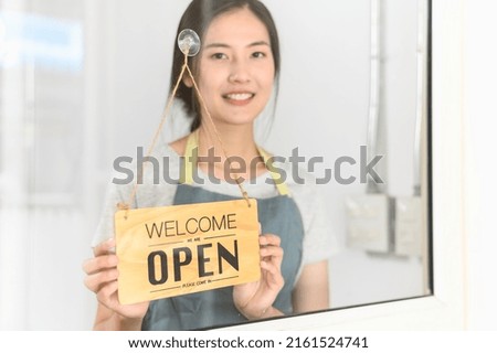 Asian small business owner woman turning shop entrance sign to open again after the quarantine due to coronavirus pandemic. Woman hanging open sign on the glass window. Focus on sign.