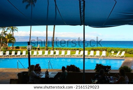 Silhouetted people sitting around dinning tables over looking a swimming  pool with the emerald blue  waters of the Pacific Ocean in the background.