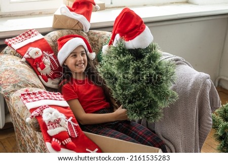Photo of cute cheerful little girl makes an artificial Christmas tree wear santa hat indoors