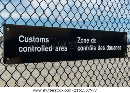A bilingual Customs Controlled Area sign in English and French on a chain link fence. Angled view with blue sky above, parking lot below.