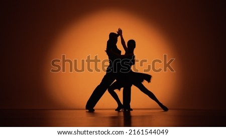 Couple of dancers approach each other and begin to dance Argentine tango. Elements of latin ballroom dance in studio with orange brown background. Dark silhouettes. Slow motion ready, 4K at 59.94fps. Royalty-Free Stock Photo #2161544049