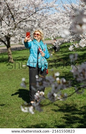 Attractive female makes a selfie with her phone in a sakura cherry tree japanese orchard. Stylish woman between flowering trees with a smartphone.