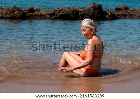 older woman sitting on the shore of the beach in orange bikini, enjoying a refreshing swim