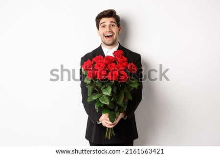 Image of handsome boyfriend in black suit, holding bouquet of red roses and smiling, being on a date, standing over white background