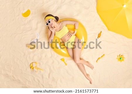 Happy relaxed brunette woman wears headband sunglasses and swimsuit chills at seaside poses on inflated swimring enjoys sunbathe on beach has cheerful expression. Summer holidays and vacation concept