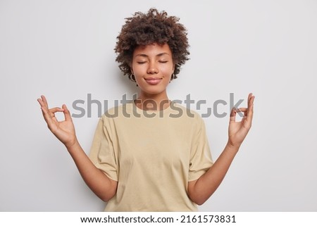 Relaxed peaceful curly haired young woman makes zen gesture keeps hads in mudra closes eyes practices yoga wears casual t shirt isolated over white background calms down during stressful day.