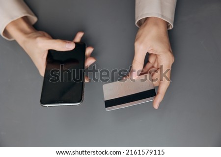 Top view on hands of tanned Latin woman in linen shirt with credit debit card and modern smartphone on dark gray background. Copy space Mockup Banner. Shopaholic lady doing online shopping.