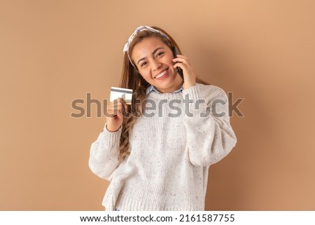 Beautiful attractive young woman posing happily and showing plastic credit card while holding mobile phone, in white sweater. Isolated over brown background.