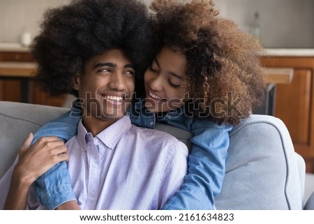 Cheerful teenage Black couple dating at home, hugging, talking, laughing, enjoying leisure. Cute teenage African girlfriend embracing, cuddling fuzzy haired boyfriend with love, affection