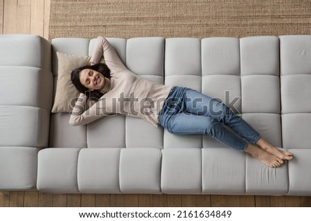 Relaxed cheerful beautiful 20s girl resting on soft comfortable big sofa, thinking, dreaming, smiling at good thoughts, planning future vacation, weekend, enjoying leisure at home, relaxing Royalty-Free Stock Photo #2161634849