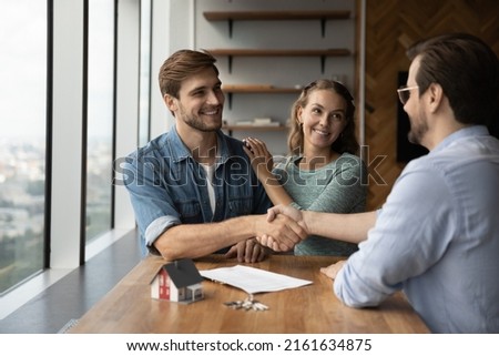 Happy couple of customers and real estate agent shaking hands. Clients and lawyer, realtor, property seller giving handshakes over negotiation table with tiny toy house model and key from new home Royalty-Free Stock Photo #2161634875