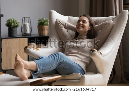 Cheerful relaxed beautiful 20s girl resting in comfortable armchair with closed eyes, peaceful smile, comfort, breathing fresh air. Smiling at good thoughts, laughing. Cozy home, relaxation concept Royalty-Free Stock Photo #2161634917