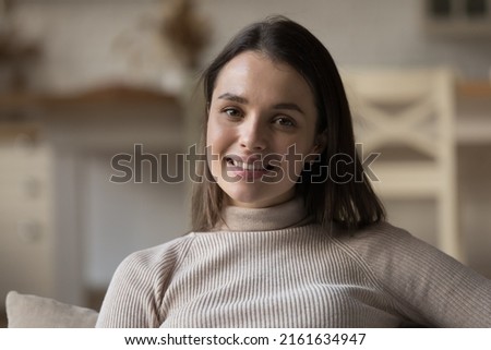 Happy millennial tenant girl resting on home sofa, looking at camera with toothy smile, posing in apartment. Renter, homeowner young woman head shot portrait. Video call screen view