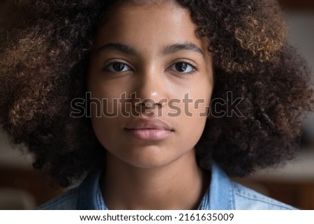 Calm serious beautiful millennial African American woman with thick curly hair looking forward at camera posing indoors. Attractive focused young adult gen z lady face without smile. Close up portrait Royalty-Free Stock Photo #2161635009