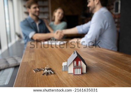 Toy house and keys with family couple of clients and real estate agent giving handshakes in background. Customers meeting with broker, property seller, lawyer, shaking hands over buying contract