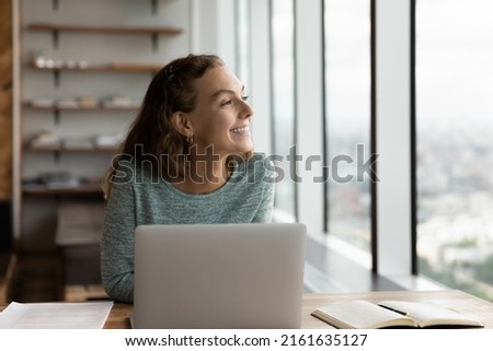 Happy student girl looking out of window and dreaming at workplace with laptop. Young employee sitting at computer in office, thinking of good future vision, smiling at positive thoughts Royalty-Free Stock Photo #2161635127