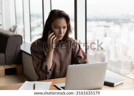 Serious businesswoman talking on mobile phone at office workplace, working at computer. Secretary answering telephone call. Manager, advisor giving consultation to customer on cellphone. Multitasking Royalty-Free Stock Photo #2161635179