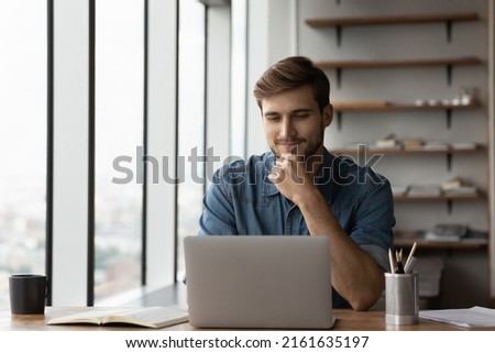 Happy satisfied millennial man using laptop at office workplace, working from home, reading message with good news, thinking, looking at screen, watching online webinar, training, making payment Royalty-Free Stock Photo #2161635197
