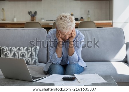 Stressed concerned aged female retiree sit on couch at home office hug head in depression unable to pay utility bills shocked with too high tax rates. Distressed older age female has financial problem Royalty-Free Stock Photo #2161635337