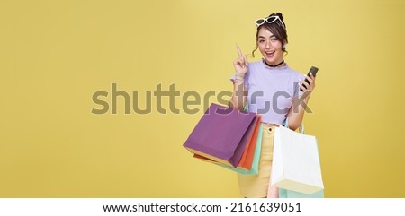 Cheerful happy teen asian woman enjoying shopping, she is carrying shopping bags and hand pointing isolated over yellow background.