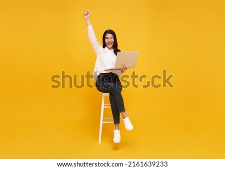 Young woman asian happy smiling celebrate. While her using laptop sitting on white chair isolate on bright yellow background.
