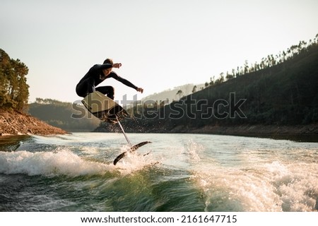 young active man in black wetsuit jump with foil wakeboard over splashing wave. Water sports activity. Royalty-Free Stock Photo #2161647715