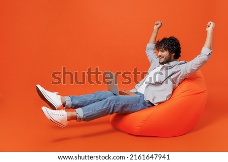 Full size young bearded Indian man 20s he wears blue shirt sit in bag chair hold use work on laptop pc computer doing winner gesture clenching fists isolated on plain orange background studio portrait