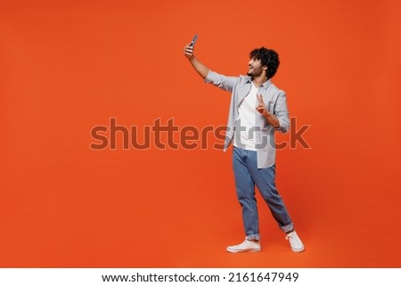 Full size body length young bearded Indian man 20s years old wears blue shirt doing selfie shot on mobile cell phone post photo on social network go isolated on plain orange background studio portrait